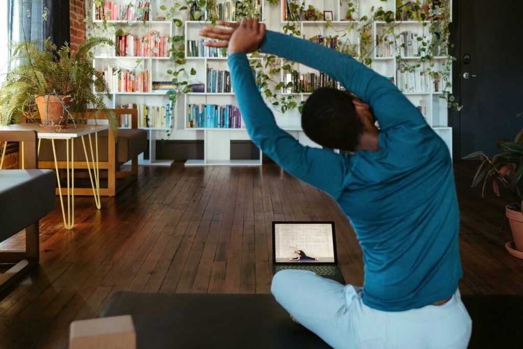 Person stretching on a mat at home in front of a laptop, with bookshelves and plants behind